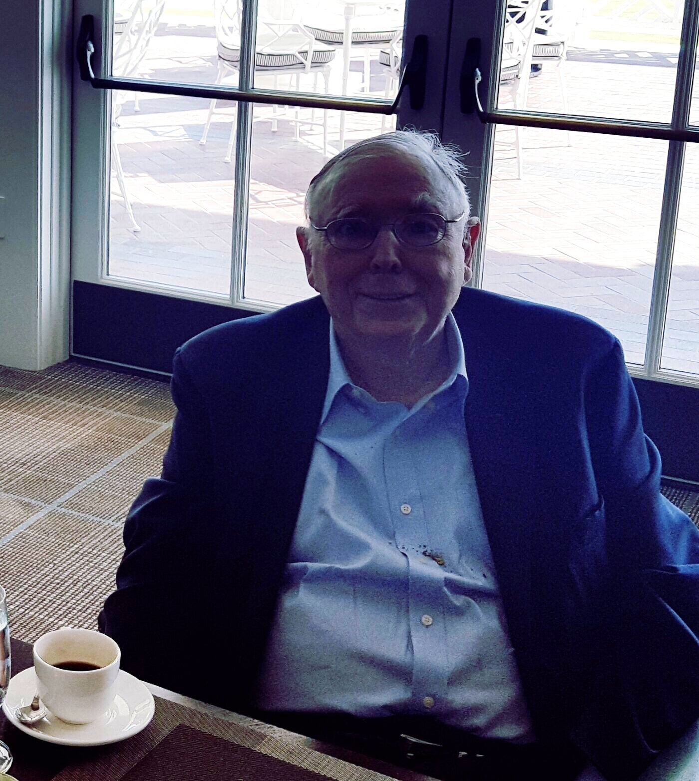 Charlie Munger at a club luncheon table in a navy blazer and open-collar shirt, a cup of coffee in front of him, the terrace and the golf course through the glass behind