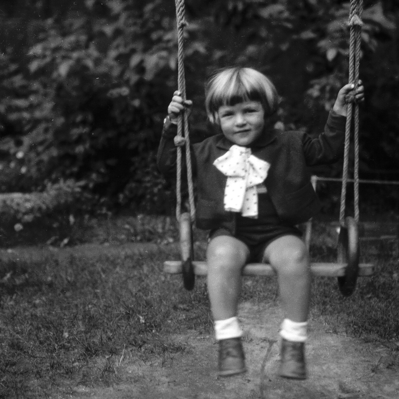 George Soros as a small boy, sitting on a rope swing in a dark jacket and a white polka-dot bow